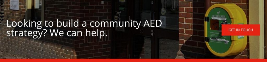 Looking to build a community AED strategy We can help: A banner with the text "Looking to build a community AED strategy? We can help." alongside a red "Get In Touch" button, set against a background photograph of a yellow defibrillator cabinet mounted on a brick wall outside a building.