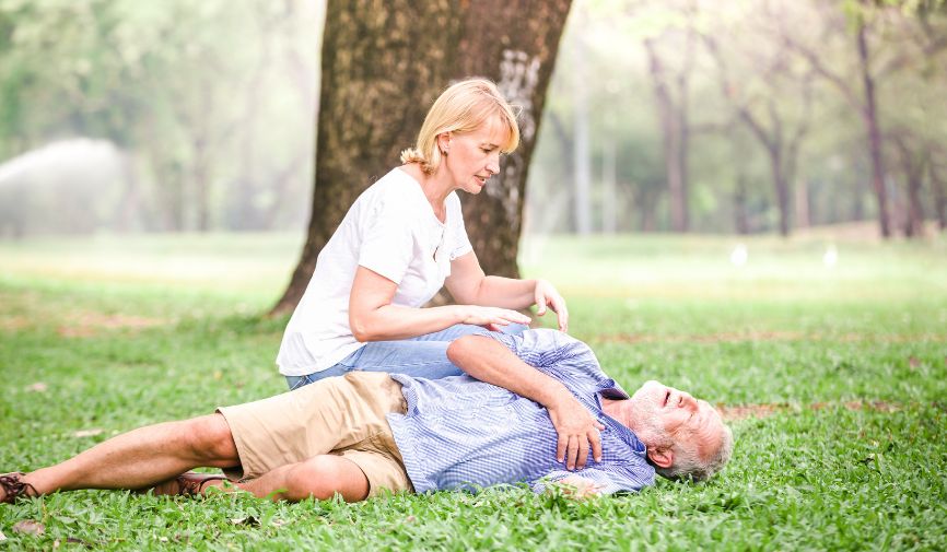 Community AED provision UK: A woman kneels beside an unconscious older man lying on grass in a park, performing a check on him with one hand on his chest.