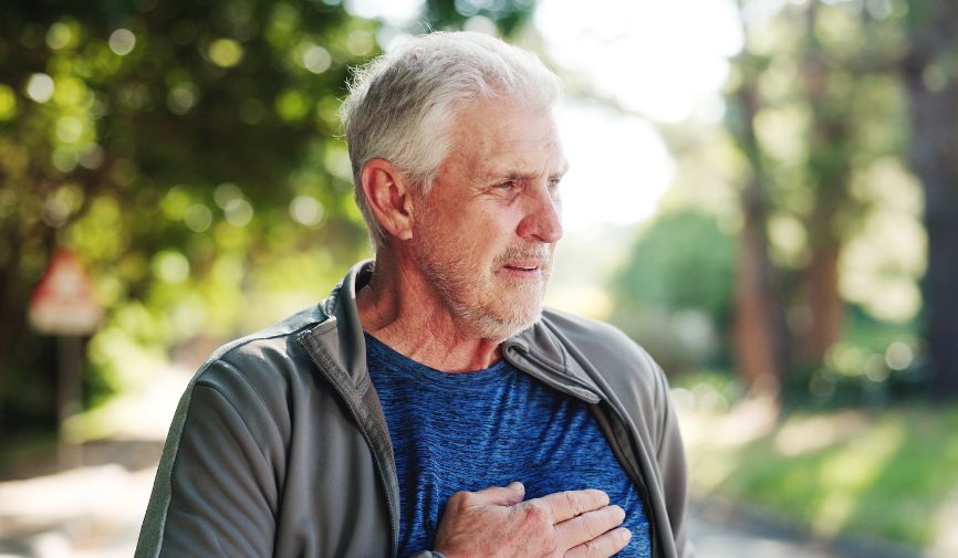 Defibrillator for community spaces: An older man outdoors in sportswear, holding his hand to his chest with a concerned expression, suggesting possible cardiac symptoms.