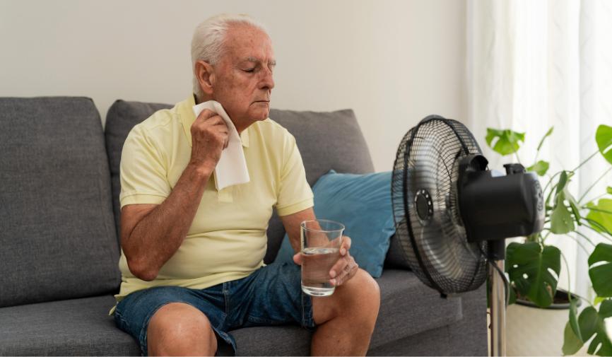 Defibrillators for workplaces An elderly man sitting on a sofa, wiping his neck with a cloth and holding a glass of water while cooling down in front of an electric fan.