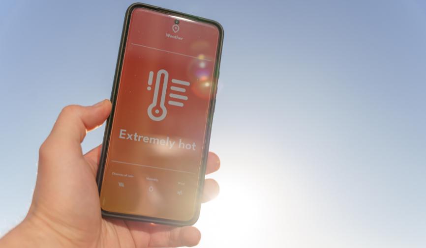 Heatwaves and heart health A hand holding a smartphone displaying a weather app with an "Extremely hot" warning against a bright, sunny sky.