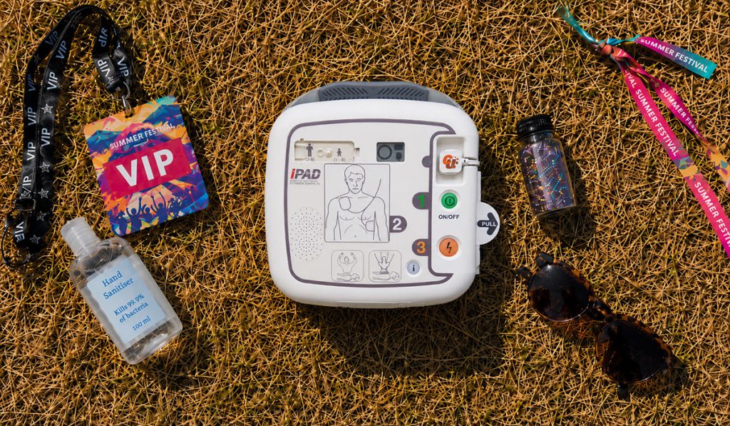 Summer Event First Aid Planning: A white iPAD defibrillator lying on dry summer grass, surrounded by a VIP festival lanyard and pass, a bottle of hand sanitiser, sunglasses and colourful festival wristbands.