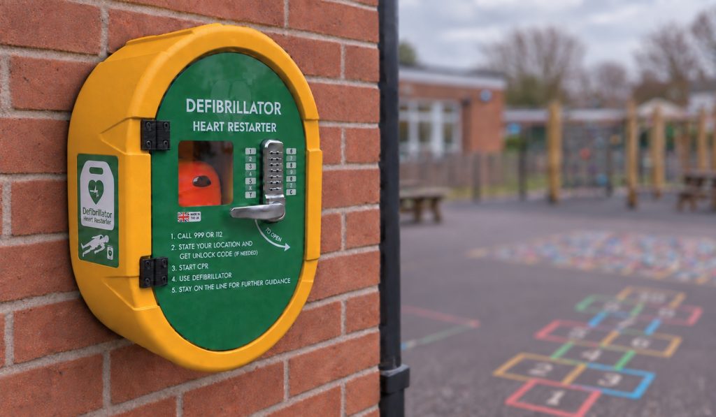 playground AED storage: A yellow and green wall-mounted defibrillator cabinet with a keypad lock fixed to a brick wall, with a school playground featuring hopscotch markings and climbing equipment in the background.
