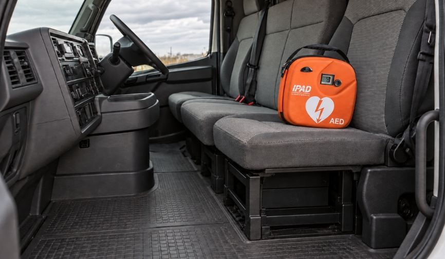 defibrillator for the workplace: An orange iPAD AED defibrillator in a carry case resting on the grey passenger seat inside the cab of a truck or commercial vehicle.