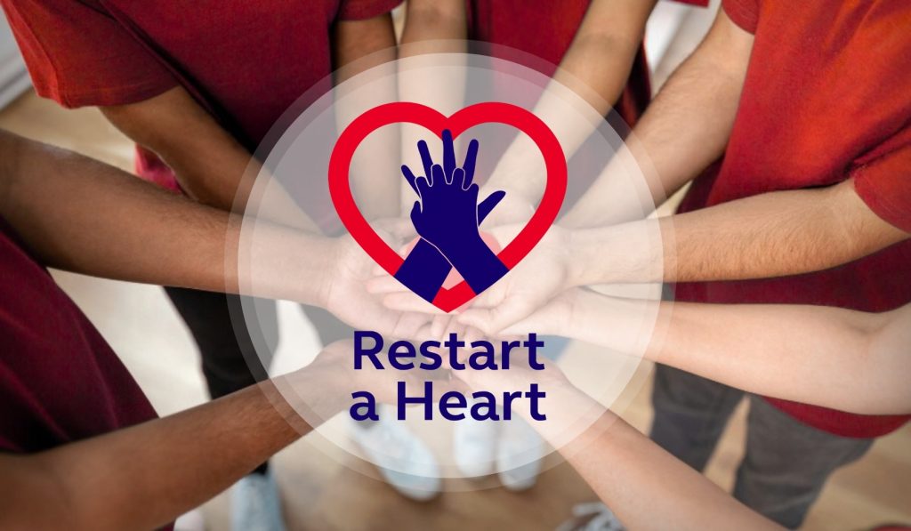 Overhead view of a group of people in red T-shirts with their arms stretched into the centre, hands stacked together. Over the centre is the Restart a Heart logo, a red heart outline with navy hands doing CPR chest compressions and the words "Restart a Heart", on a soft white circle.